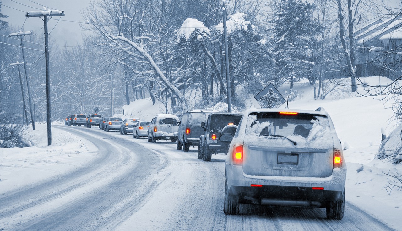 A line of cars slowly driving on a snowy, icy road. Entire image is monochrome blue-ish except the taillights, which are glowing red and yellow.