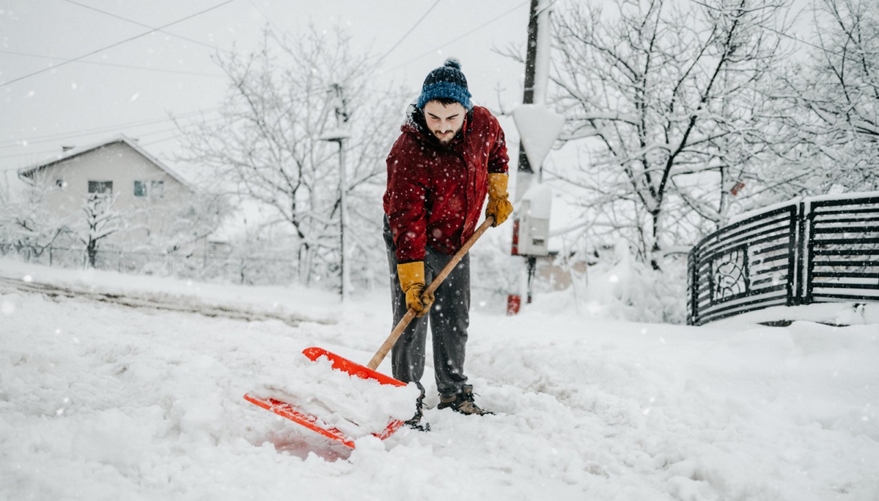 Man in red jacket with snow shovel cleans sidewalks and city street in winter. Cloudy winter day.